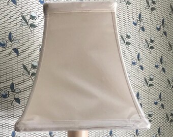 May include: A white lampshade with a square shape and a slightly curved bottom edge. The lampshade is made of a fabric material and has a smooth, even texture.
