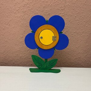 Könnte beinhalten: Eine blaue Holzblume mit gelber Mitte und einer kleinen Holztür. Die Blume hat grüne Blätter und einen grünen Sockel.