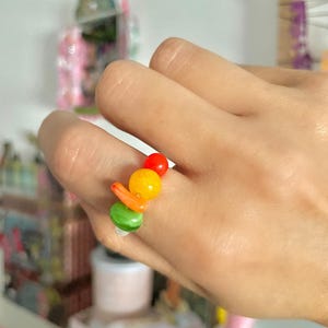 Può includere: Un anello di perline colorate con perline rosse, gialle, verdi e arancioni. L'anello è indossato su un dito, mostrando la disposizione giocosa delle perline. Le perline sono rotonde e oblunghe.