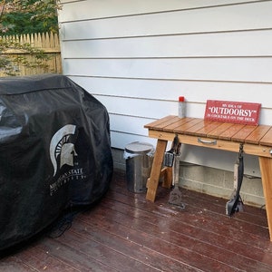 Pu&ograve; includere: Una copertura per barbecue nera con il logo della Michigan State University, un tavolo di legno con un cartello che recita "My idea of "OUTDOORSY" is cocktails on the deck", e un bidone della spazzatura in metallo.