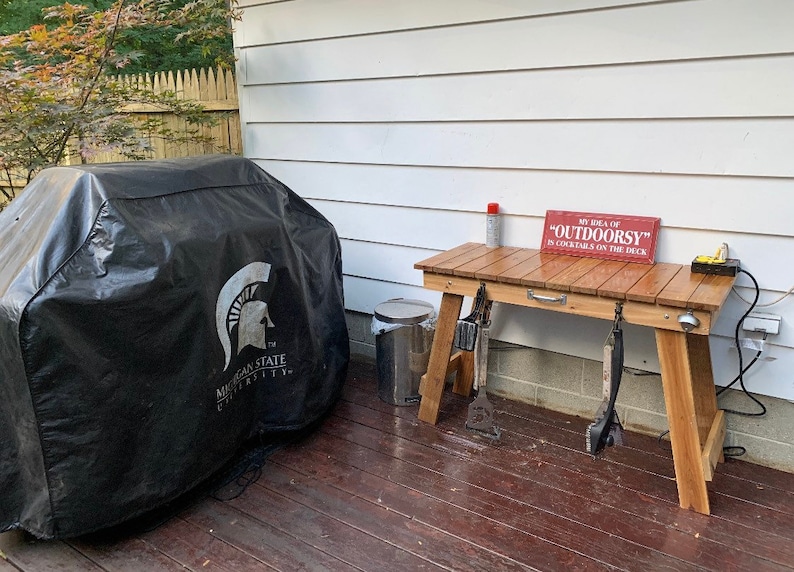Pu&ograve; includere: Una copertura per barbecue nera con il logo della Michigan State University, un tavolo di legno con un cartello che recita "My idea of "OUTDOORSY" is cocktails on the deck", e un bidone della spazzatura in metallo.
