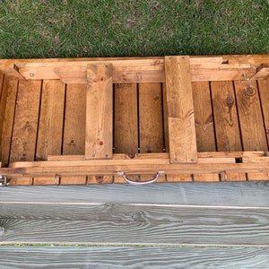 Pu&ograve; includere: Un tavolo pieghevole in legno con una maniglia e ganci sul fondo. Il tavolo &egrave; in legno naturale e ha una macchia marrone chiaro.