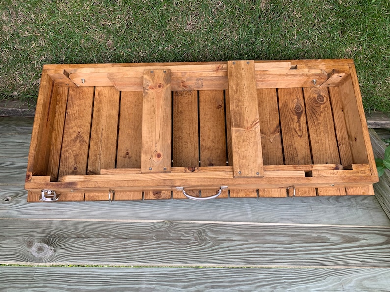 Pu&ograve; includere: Un tavolo pieghevole in legno con una maniglia e ganci sul fondo. Il tavolo &egrave; in legno naturale e ha una macchia marrone chiaro.