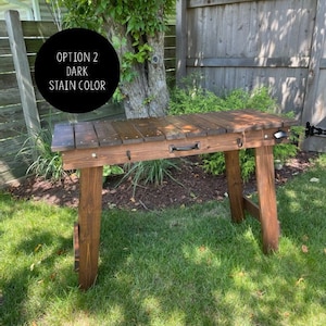 Pu&ograve; includere: Un tavolo in legno scuro con un aspetto rustico. Il tavolo ha un unico cassetto con una maniglia nera ed &egrave; sostenuto da due gambe a forma di A. Il testo "OPTION 2 DARK STAIN COLOR" &egrave; visibile nell'immagine.