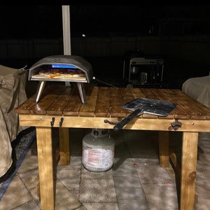 Pu&ograve; includere: Un tavolo di legno con un forno per pizza, una pala per pizza e una bombola di propano. Il tavolo &egrave; in legno naturale e ha un aspetto rustico. Il forno per pizza &egrave; nero e ha una porta di vetro. La pala per pizza &egrave; in metallo e ha un manico di legno. La bombola di propano &egrave; bianca e ha un'etichetta rossa.