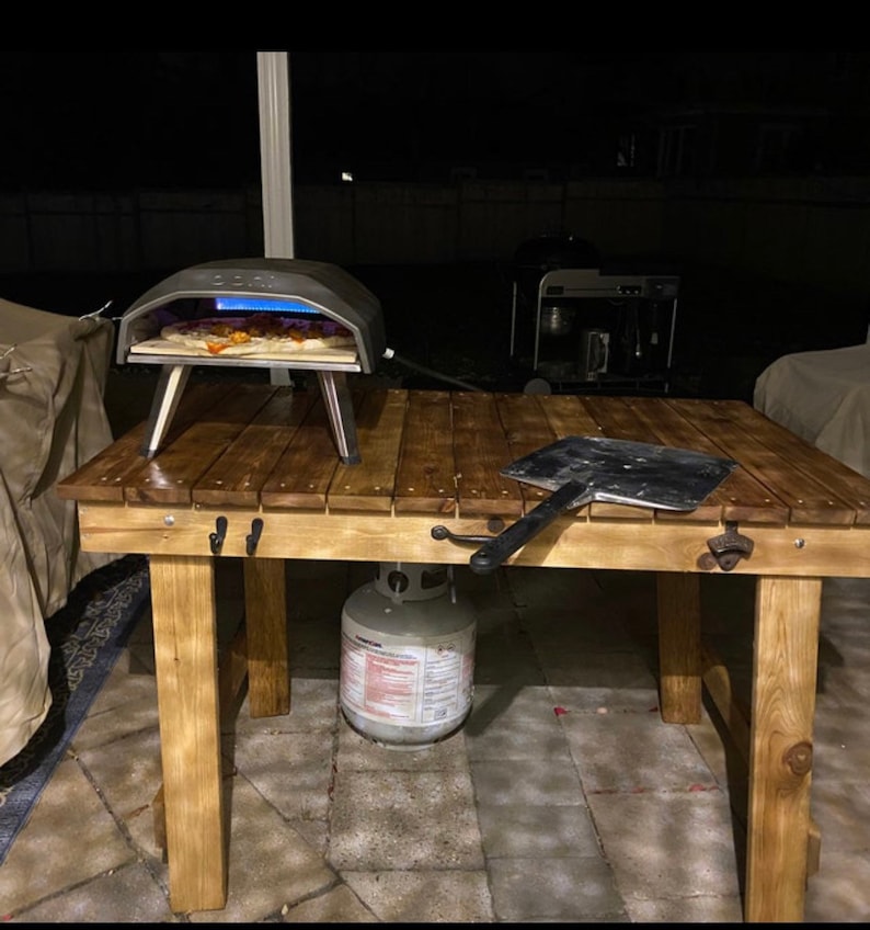 Pu&ograve; includere: Un tavolo di legno con un forno per pizza, una pala per pizza e una bombola di propano. Il tavolo &egrave; in legno naturale e ha un aspetto rustico. Il forno per pizza &egrave; nero e ha una porta di vetro. La pala per pizza &egrave; in metallo e ha un manico di legno. La bombola di propano &egrave; bianca e ha un'etichetta rossa.