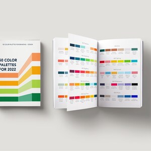 Op de afbeelding: Een gesloten boek met de titel "50 Color Palettes for 2022" met een kleurrijk grafisch ontwerp op de omslag. Het boek is open op een pagina met een raster van kleurpaletten, elk met een naam en een set kleursamples.