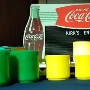 May include: A collection of green and yellow mugs. The mugs are arranged in a row on a dark blue surface. A Coca-Cola sign is visible in the background.