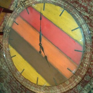 May include: A round wooden clock with a glass face. The clock face is painted with colorful stripes in shades of red, orange, yellow, and brown. The clock hands are black and point to the time of 3:45.