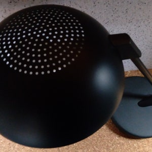May include: A black desk lamp with a round, perforated shade and a black base. The lamp has a flexible arm and a black cord.