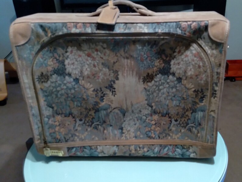 May include: A vintage suitcase with a floral tapestry design. The suitcase is brown with a tan leather trim and a handle.