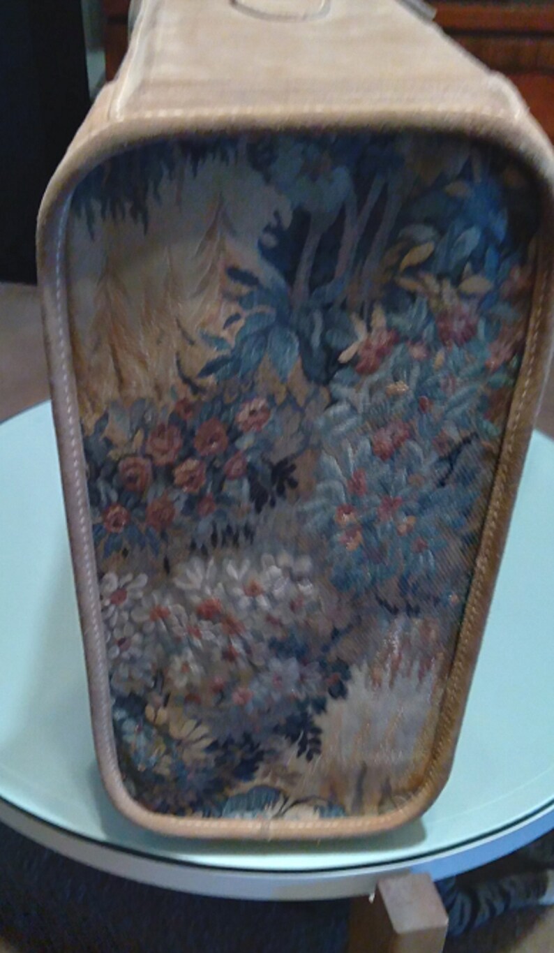 May include: A beige vintage suitcase with a floral tapestry design. The tapestry features a variety of flowers, leaves, and trees in shades of blue, green, red, and white.