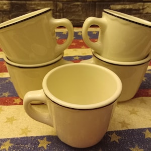May include: Four white ceramic mugs with black trim stacked on top of each other. The mugs are on a red, white, and blue patterned surface.