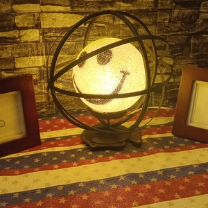 May include: A black metal globe lamp with a white smiley face light bulb. The lamp is on a table with two brown picture frames. The table has a red, white, and blue patterned tablecloth.
