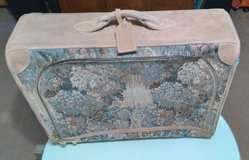 May include: A vintage floral tapestry suitcase with a tan leather trim and handle. The suitcase has a zipper closure and a luggage tag.