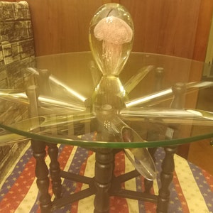 May include: A glass coffee table with a clear, round top supported by a black wooden base. The table features a central glass sculpture of a jellyfish with pink details. The table's legs extend outwards, creating a unique design.