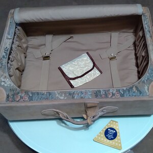 May include: A vintage floral patterned suitcase with a brown leather trim and a beige interior. The suitcase is open and has a small beige pouch inside.