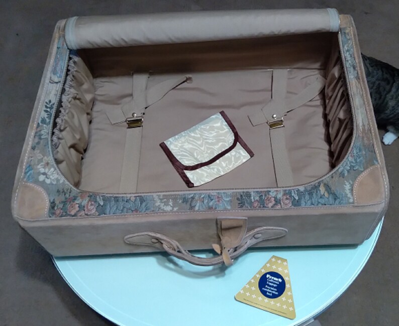 May include: A vintage floral patterned suitcase with a brown leather trim and a beige interior. The suitcase is open and has a small beige pouch inside.