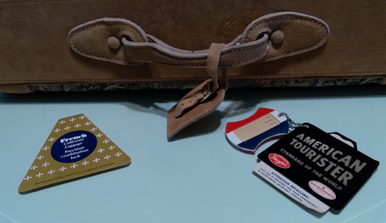 May include: A brown leather suitcase with a floral pattern. The suitcase has a leather handle and a metal latch. There are three tags attached to the suitcase, one with the text "American Tourist Standard of the World", one with the text "Preach California Luggage Precision Combination Lock", and one with a blank space for writing.
