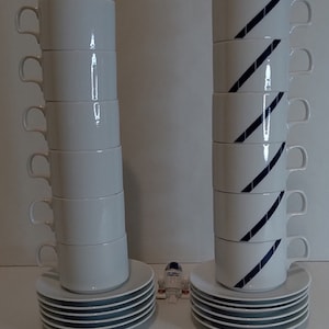 May include: Two stacks of white ceramic coffee mugs with blue stripes. The mugs are stacked on top of each other and are on white saucers. There are eight mugs in each stack.