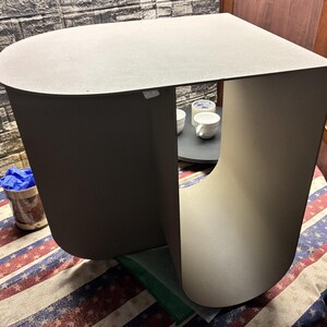 May include: A matte gray side table featuring a continuous, curved C-shaped design. The structure is formed from a single piece of material with a flat top surface and rounded edges, creating an open storage space underneath the tabletop.