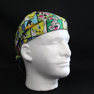 May include: A colorful surgical scrub cap featuring a repeating pattern of cartoon characters, including Bugs Bunny, Daffy Duck, and Tweety Bird. The cap is primarily green, blue, and yellow, and is displayed on a white mannequin head against a black background.