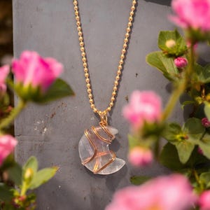 May include: A gold-colored chain necklace with a crescent moon pendant. The pendant is a light-colored stone wrapped in copper wire. Pink flowers are in the foreground, and the background is a gray surface.