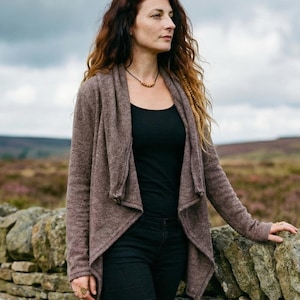 May include: A woman in a brown cardigan, black top, and trousers stands by a stone wall. She wears brown boots and a necklace. The background shows a field of purple flowers under a cloudy sky.