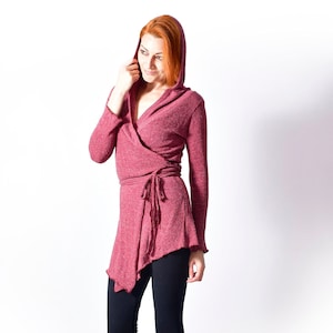 May include: A woman wearing a pink hooded wrap cardigan with a tie at the waist and black leggings. She is wearing black ankle boots and standing on a woven rug.
