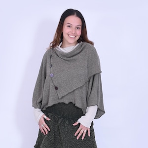 Könnte beinhalten: Ein olivgrünes Ensemble mit einem mehrschichtigen Rock und einem drapierten Poncho-Oberteil. Der Rock hat ein mehrschichtiges Design mit strukturiertem Stoff, und das Oberteil wird mit Knöpfen befestigt. Das Outfit wird mit neutralfarbenen Sandalen mit Absatz kombiniert.