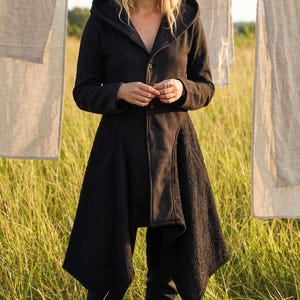 May include: A black hooded coat with an unusual asymmetrical design. The coat has a button-up front and a flared skirt-like hem. The model is in a field, wearing dark trousers and brown boots.