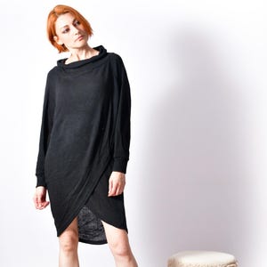 May include: A black long-sleeved dress with a cowl neck and wrap-style skirt. The dress is worn with black ankle boots. A small, round stool with a cream top and a black and white patterned base is also visible.