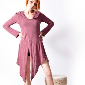 May include: A mauve hooded dress with long sleeves and an asymmetrical hemline. The dress is worn with black ankle boots. The model is standing on a woven rug in front of a white background.