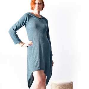 以下が含まれることがあります： 長袖と非対称の裾が付いた、ティール色のフード付きドレス。ドレスは黒のアンクルブーツと合わせています。クリーム色のトップと白黒の模様のバンドが付いた、小さくて丸いスツールが近くにあります。