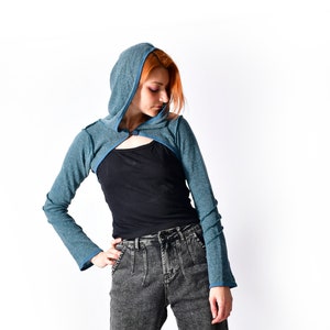 May include: A teal blue hooded crop top with long sleeves and a button closure. The top is worn over a black tank top and dark gray jeans.