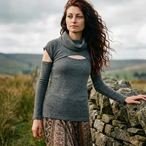 May include: A grey long-sleeved top with a keyhole cutout and a matching cowl neck. The top is paired with a patterned skirt. The model is standing next to a stone wall in a natural outdoor setting.