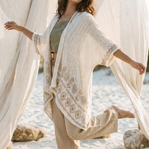 Op de afbeelding: Een vrouw draagt een crèmekleurige, open kimono met geometrisch borduurwerk, gecombineerd met een groene top en een beige broek. De outfit is licht en luchtig, perfect voor het strand of een resort. De vrouw lacht en lijkt zich te vermaken.