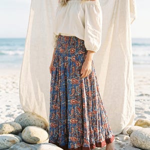 May include: A woman wearing a white off-the-shoulder top and a long, flowing skirt. The skirt features a blue paisley pattern with red and brown accents. The scene is set on a sandy beach with the ocean in the background.
