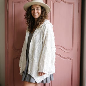 Puede incluir: Una chaqueta con capucha de color crema con un acabado de tela texturizada y deshilachada con un tejido de patrón de diamantes, sobre un vestido corto gris oscuro, con un sombrero de fieltro beige de ala ancha y botas marrón claro.