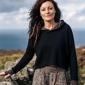 May include: A person wearing a black hoodie with patterned details and a patterned skirt, standing outdoors. The image shows a stone wall, the sea, and a cloudy sky. The clothing is casual and fashionable.