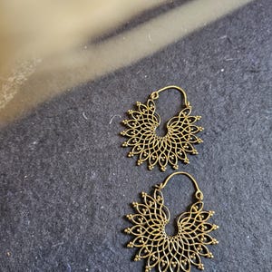 May include: A pair of gold-toned hoop earrings with a delicate, intricate design. The earrings are made of a circular, openwork pattern with a floral motif.