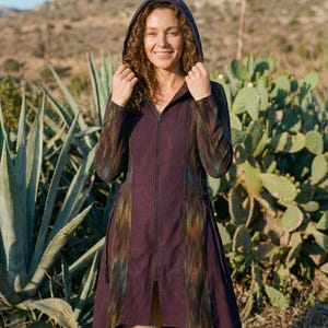 Può includere: Un cappotto con cappuccio e maniche lunghe di colore viola scuro. Il cappotto presenta una cerniera a tutta lunghezza e pannelli verticali con un design strutturato e colorato. La modella indossa pantaloni beige. Lo sfondo comprende piante del deserto e un cielo soleggiato.