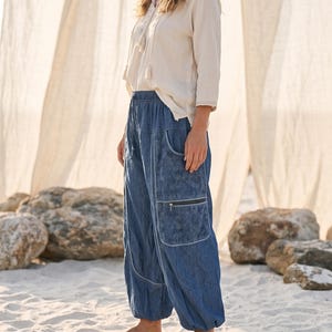 May include: A beige long-sleeved tunic with a tassel drawstring neckline paired with loose-fitting blue denim trousers featuring an elastic waistband, side pockets with floral embroidery, and a horizontal zippered pocket on the right thigh.