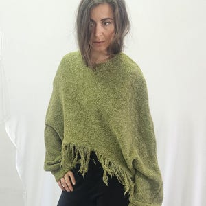 May include: A green, textured sweater with a fringed hemline. The sweater has long sleeves and a relaxed fit. The model is wearing black trousers. The background is a plain white sheet.