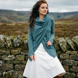 May include: A woman in a teal wrap-around sweater and white denim skirt stands by a stone wall. The sweater has a cowl neck and long sleeves. She wears brown boots. The background shows a cloudy sky and a field. The sweater is approximately 150 cm long.