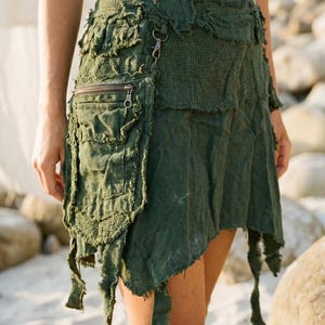 Könnte beinhalten: Dunkelgrüner Rock mit asymmetrischem, geschichtetem Design. Er hat eine Vordertasche mit Reißverschluss und Metallbeschlägen. Der Stoff hat ein abgenutztes, strukturiertes Aussehen mit ausgefransten Kanten und dekorativen Knöpfen.