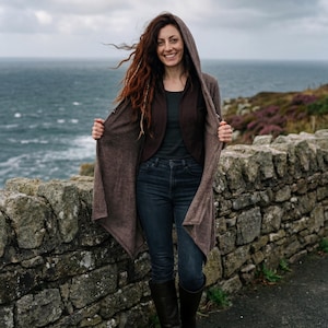 Peut inclure: Une femme porte un long cardigan marron à capuche sur un haut foncé et un jean bleu, debout près d'un mur en pierre avec l'océan en arrière-plan. Elle porte également des bottes noires. Le cardigan est ouvert, révélant la tenue en dessous.