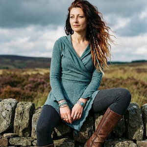 Può includere: Una persona indossa un top avvolgente color turchese con cappuccio, leggings grigi e stivali di pelle marrone seduta su un muro di pietra. Braccialetti turchesi adornano i suoi polsi. Lo sfondo mostra un campo e un cielo nuvoloso.