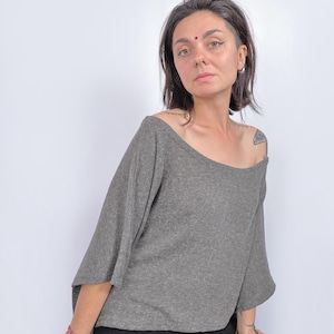Peut inclure: Un haut gris à épaules dénudées avec des manches larges. Le haut est associé à un pantalon noir. Le tissu semble être un tricot doux. Le modèle porte des bracelets et a des tatouages sur le bras.