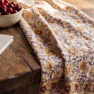 Può includere: Runner da tavolo floreale color crema con motivo floreale giallo e bordeaux. Il runner ha un bordo con frange ed è drappeggiato su un tavolo di legno. Una ciotola di ciliegie e una bottiglia di vetro sono anche sul tavolo.